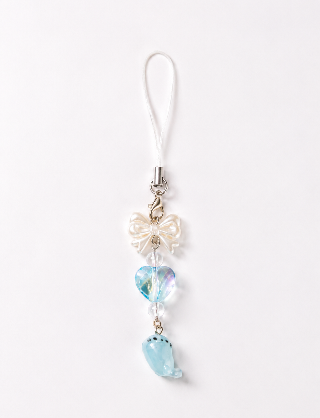 A decorative phone strap charm featuring a white loop strap, a white pearl bow, a light blue heart-shaped bead with an iridescent finish, and a light blue seal charm, set against a plain white background.