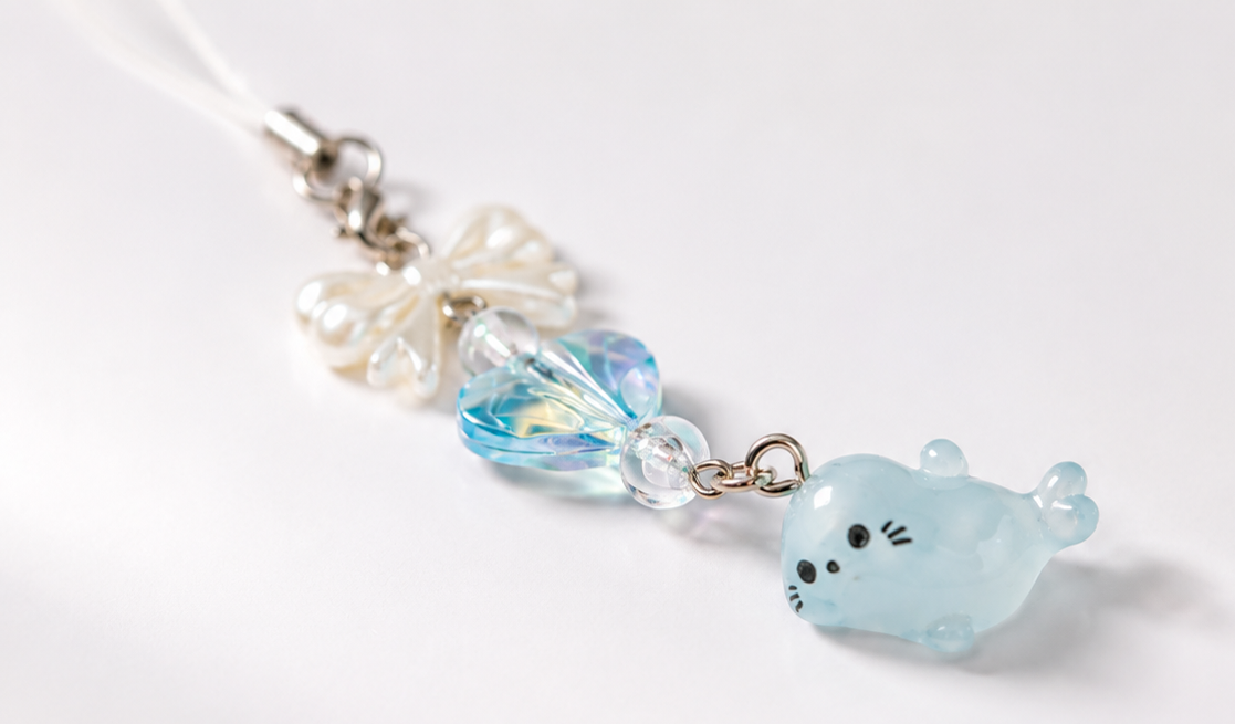 A close-up of a charm featuring a light blue translucent seal, a faceted light blue heart bead, clear beads, and a white bow on a silver chain, set against a plain white background.